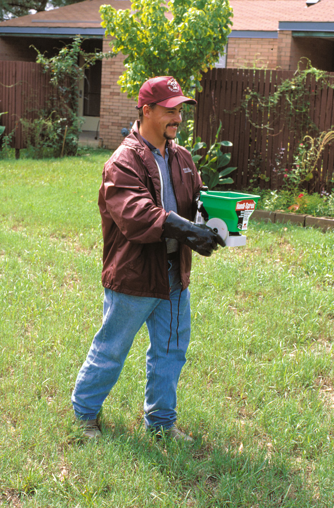 A man in a red coat and cap uses a plastic hand-operated spreader to apply an ant treatment to a yard