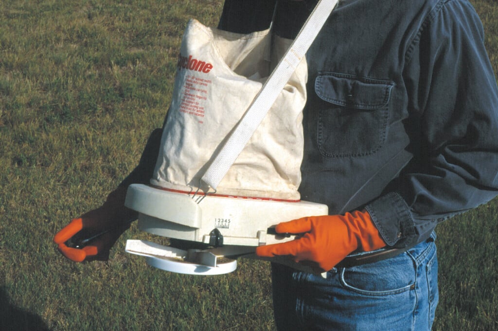 A person holds a hand-operated spreader with a bag of ant treatment attached to it