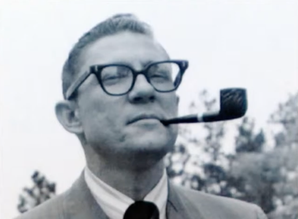 Black-and-white photo of Gale Buchanan wearing glasses and a suit, holding a pipe in his mouth, while standing outdoors.