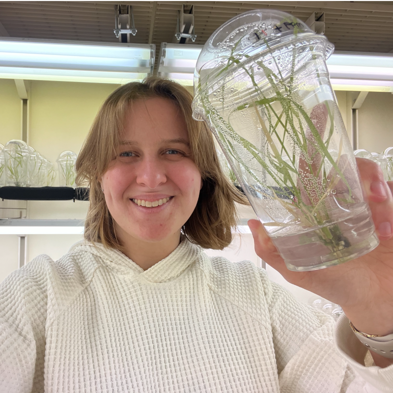 Skye Remko smiles as she holds a transparent cup with switchgrass inside in a lab setting.