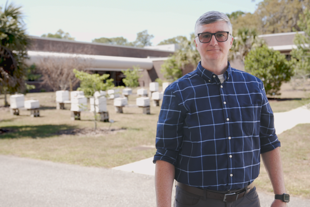 A man in a blue palid shirt stands with apiaries in the background