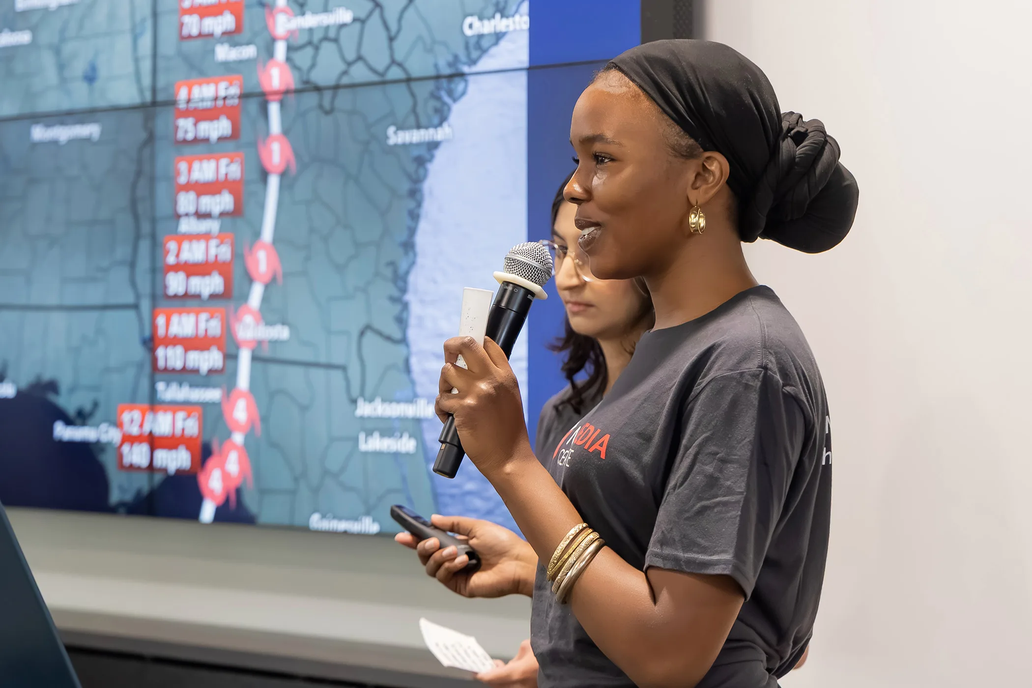 Two women stand in front of a projector showing the path of Hurricane Helene through Georgia as one woman holds a microphone and clicker.