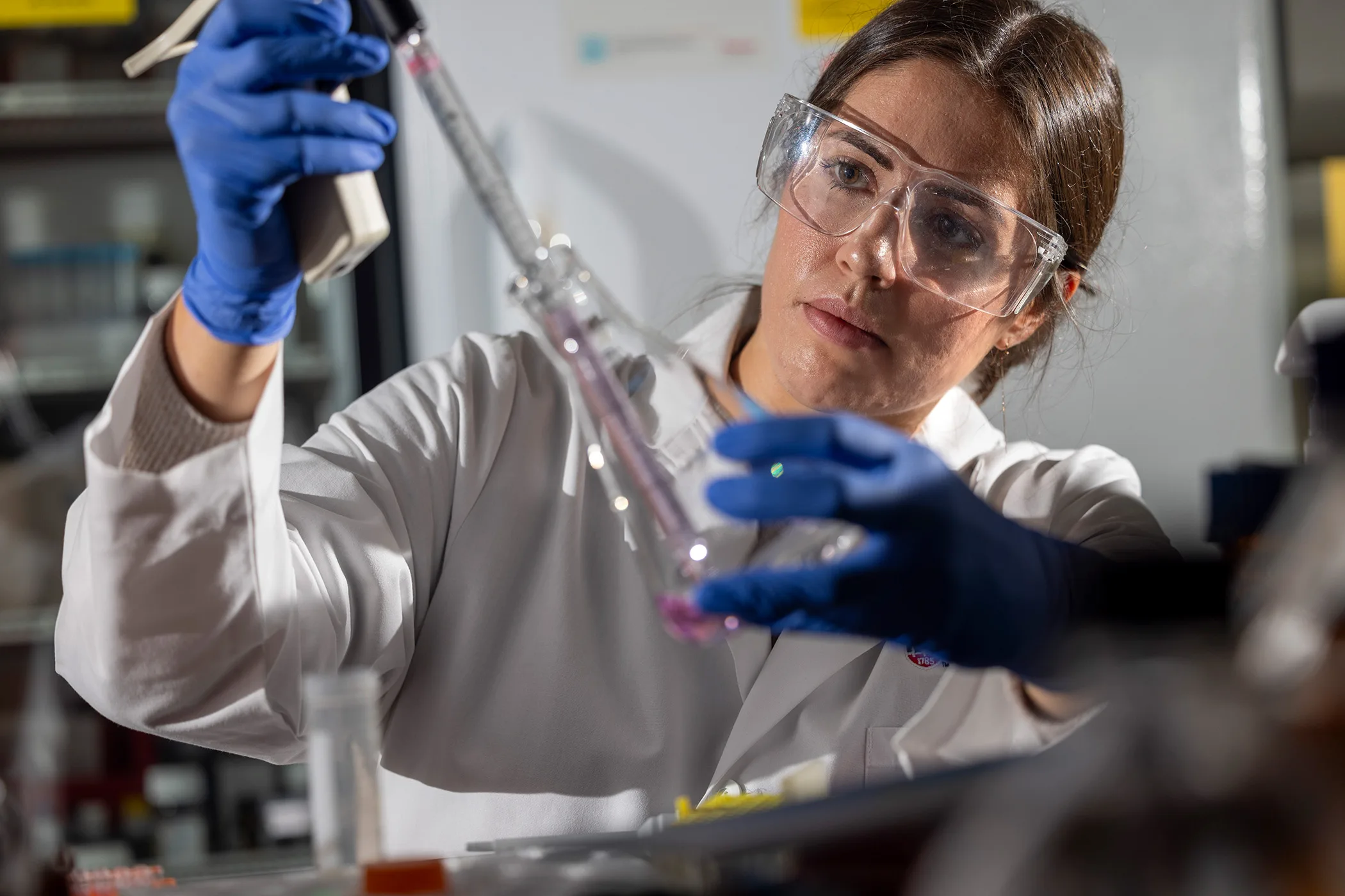 Woman wearing lab personal protective gear holds a pipette in one gloved hand and a conical flask in the other.