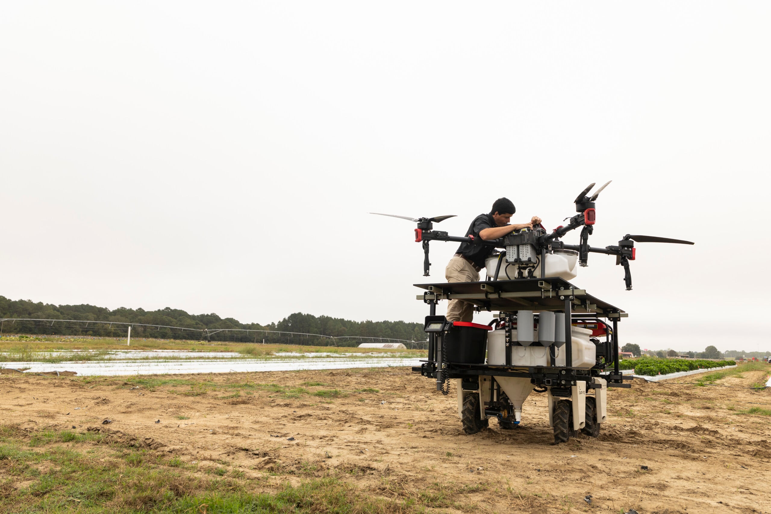 A person leans over a large cart in dirt field. On the cart is a large drone used for precise agriculture application.