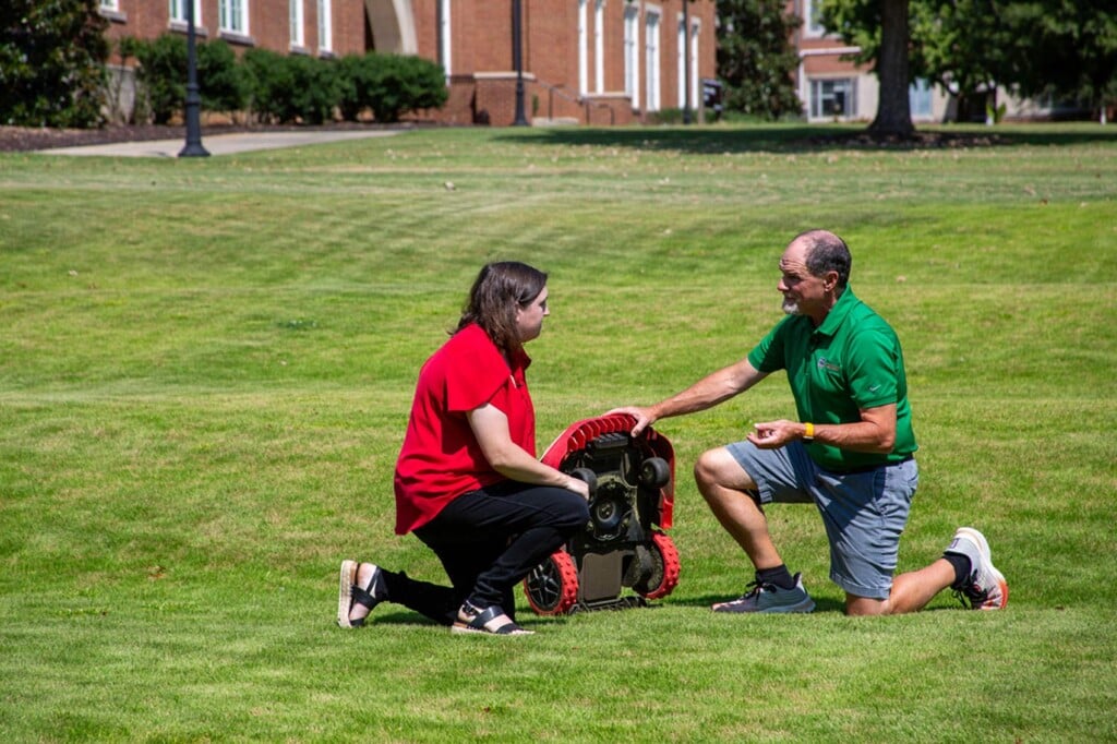 Bethany Harris and Clint Waltz kneel on a manicured lawn and examine the underside of a small autonomous mower