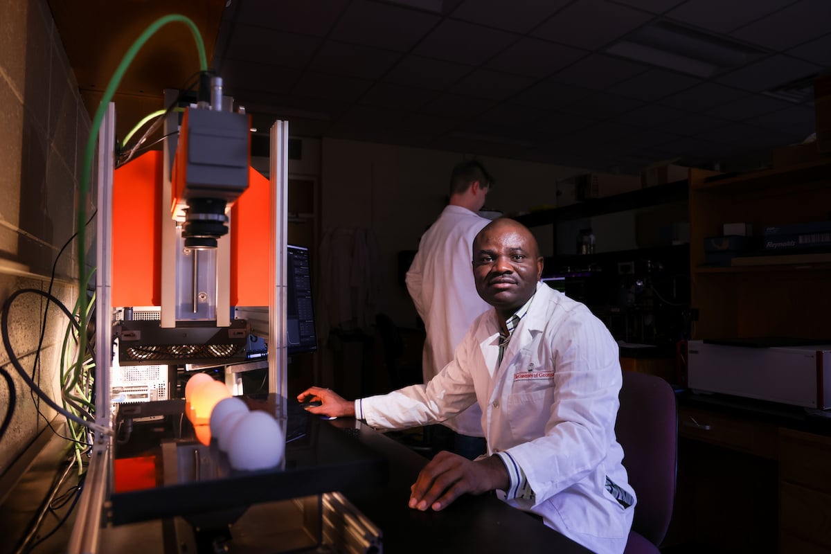 Christopher Kucha sits in a dark room at a desk with a device that projects light onto eggs. A lab assistant works in the background