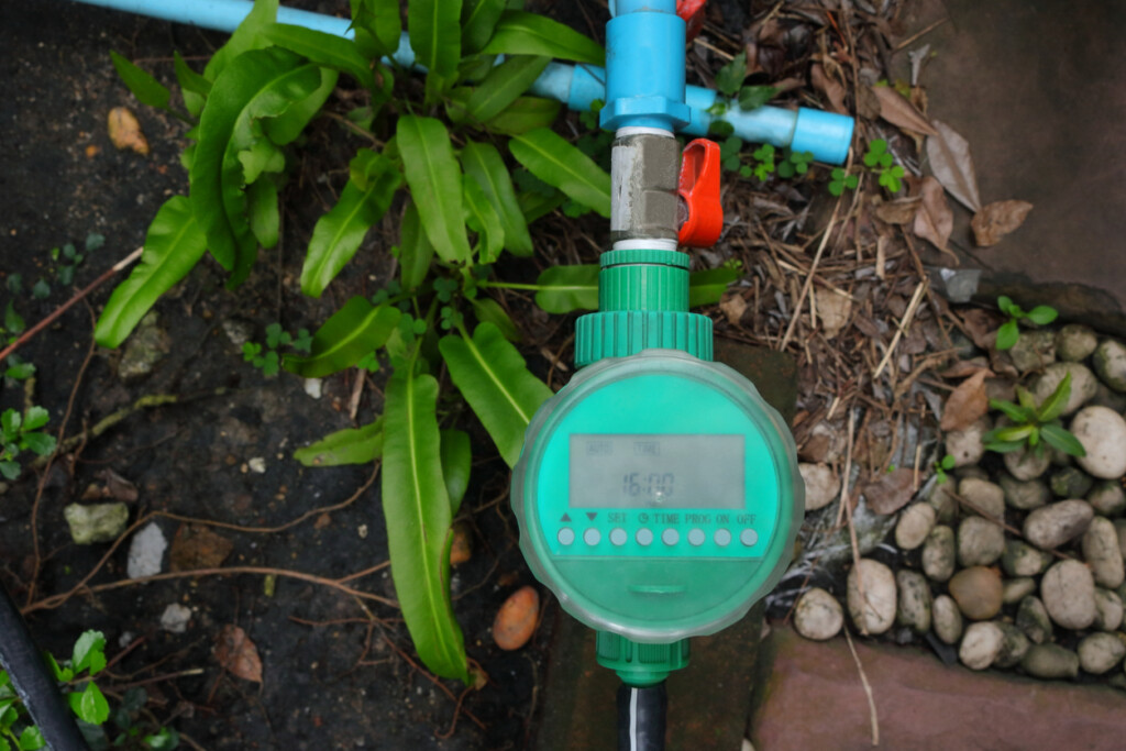 A water timer connected to irrigation pipes with soil, plants and rocks on the ground underneath.