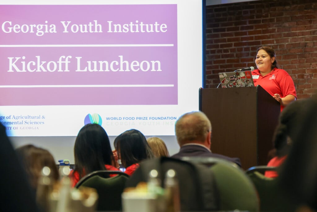 Jaky Cervantes Childers stands at a lectern with a projector in the background for the Georgia Youth Institute Kickoff Luncheon, while she addresses a room full of people looking at her. 
