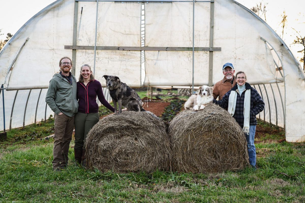 Two couples, each a man and a woman, stand in front of a hoop house next to round haybales. A dog sits on top of each haybale.