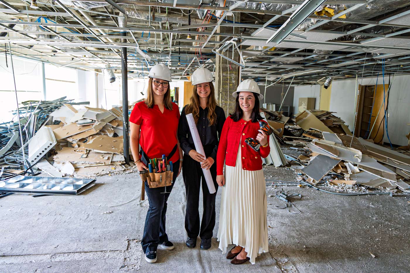 Three students in hard hats, holding tools and blueprints stand in large, demoed space in the Georgia Center
