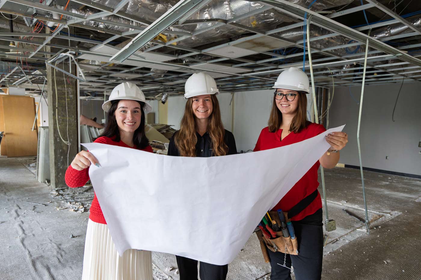 Three students in hard hats hold a blueprint in an indoor construction zone with visible duct work and ceiling framing