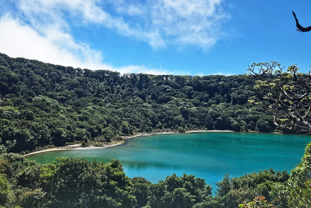 Turquoise crater lake surrounded by dense tropical forest under a bright blue sky.