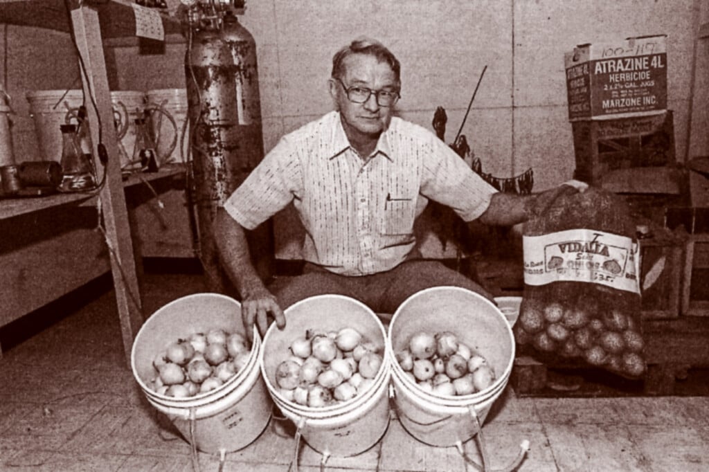 Vintage photo of Doyle Smittle crouched next to three buckets of onions with tubing attached to the buckets. He is holding a large bag of onions and behind him are a box of Atrazine herbicide, gas canisters and more buckets.