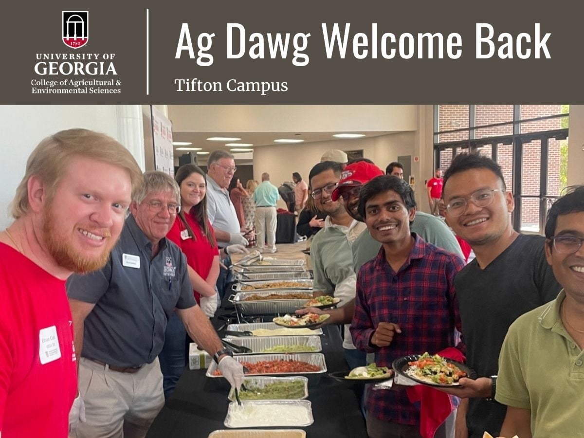 Alt text: Promotional banner for the University of Georgia College of Agricultural and Environmental Sciences titled “Ag Dawg Welcome Back – Tifton Campus.” The image shows a welcome-back meal event inside a campus building, with attendees moving through a buffet line. Serving trays of food are arranged down the center of a long table, while several students and staff stand on either side holding plates. Large windows and additional attendees are visible in the background. The UGA CAES logo appears in the upper left corner.