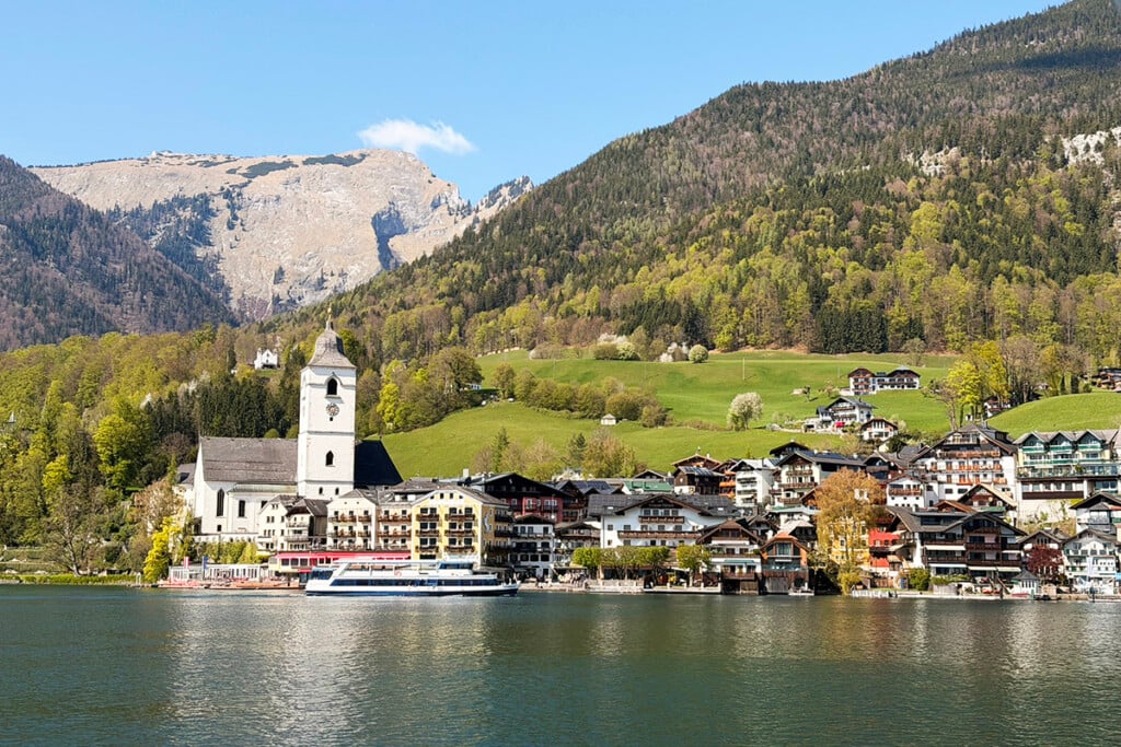 Lakeside village with a church, alpine homes, and forested mountains reflected in calm water.