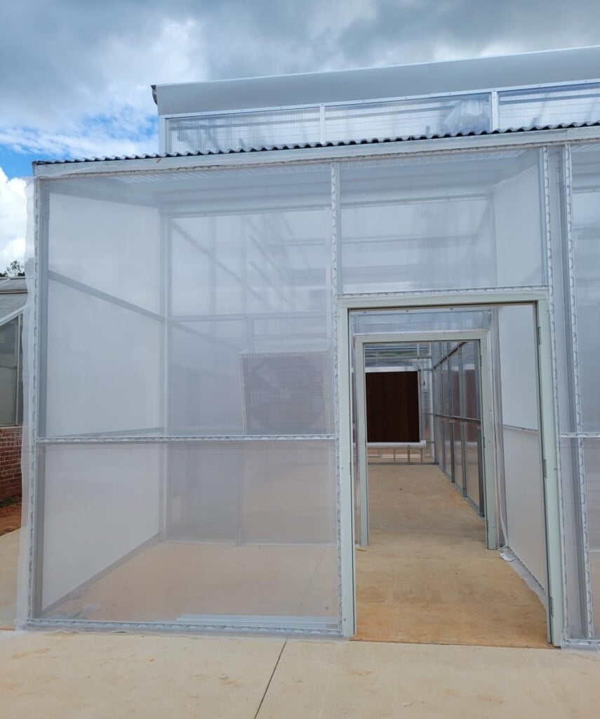 Exterior view of a large screened agricultural enclosure with a metal frame and fine mesh walls. An open doorway leads through a series of connected screened compartments with concrete floors.