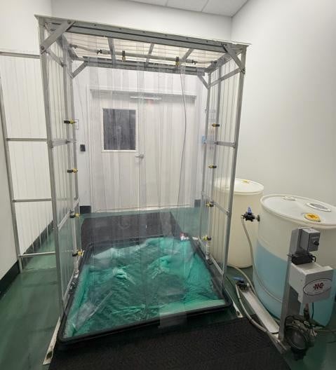 Indoor research setup with a tall rectangular metal frame enclosed by clear plastic curtains over a shallow collection basin lined with green material.