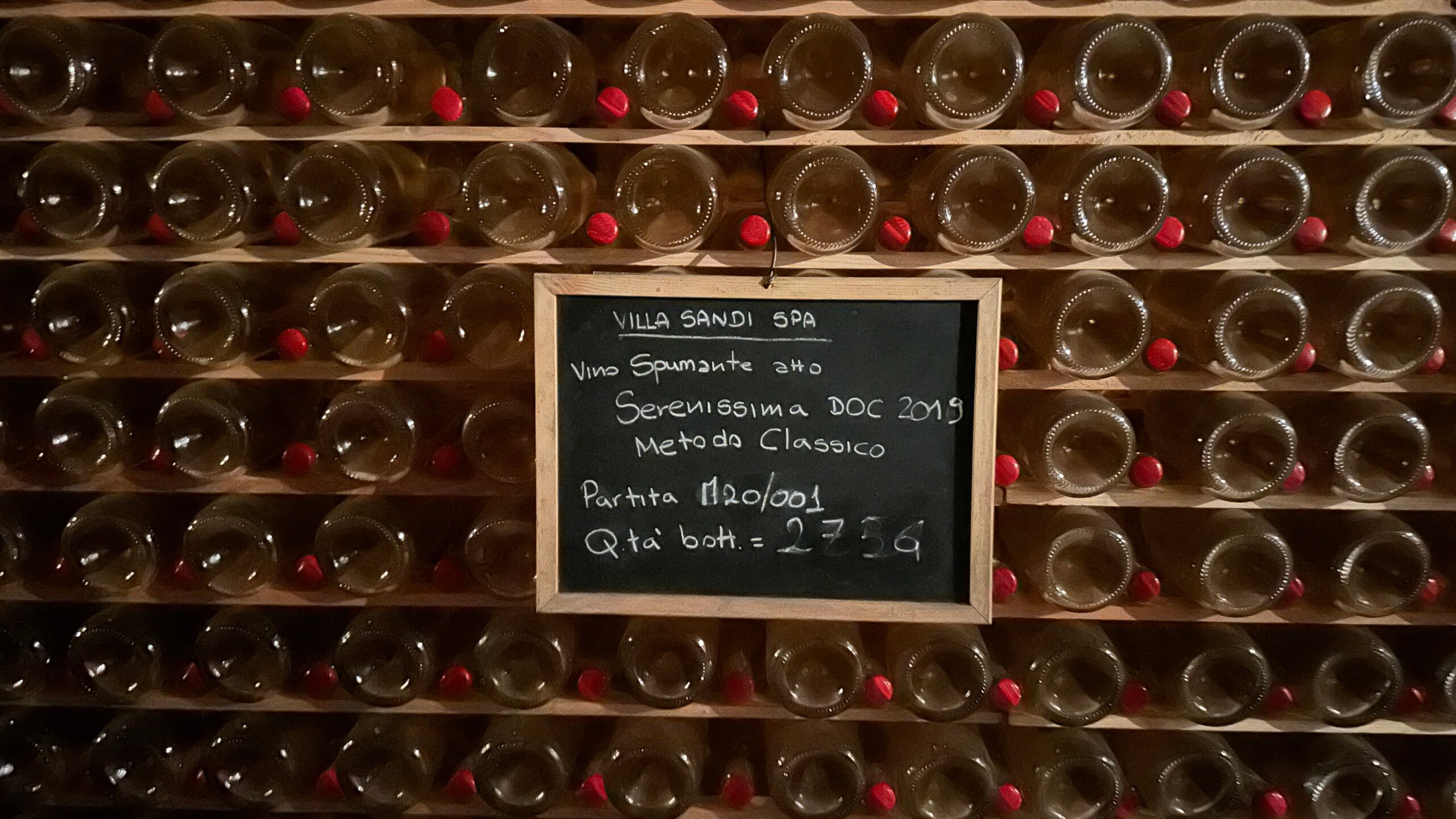 Shelves of aging sparkling wine with a chalk board with information written in Italian.