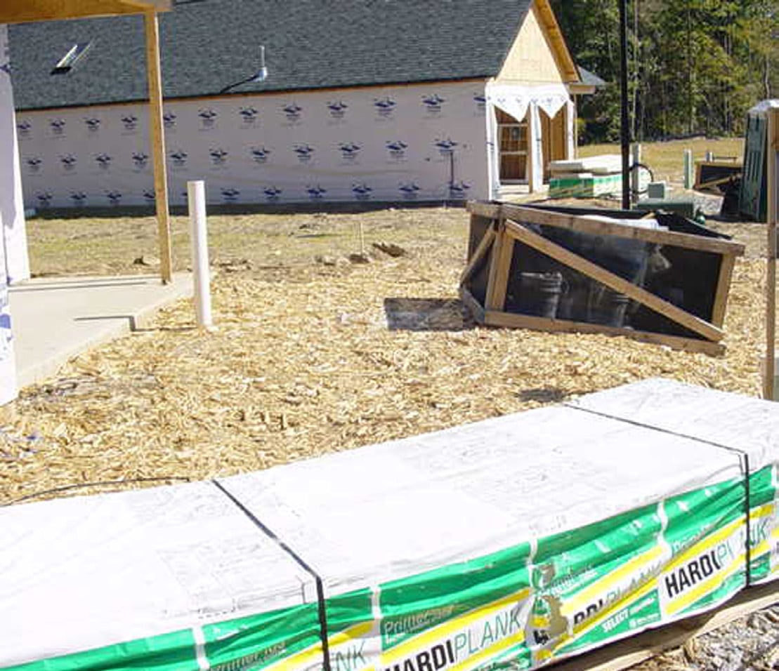 Ground wood waste spread as a mulch delivery pad in front of a house under construction, with bundled siding materials in the foreground.