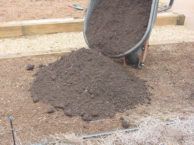 A wheelbarrow pouring dark compost or soil onto the ground next to a wood-edged garden bed.