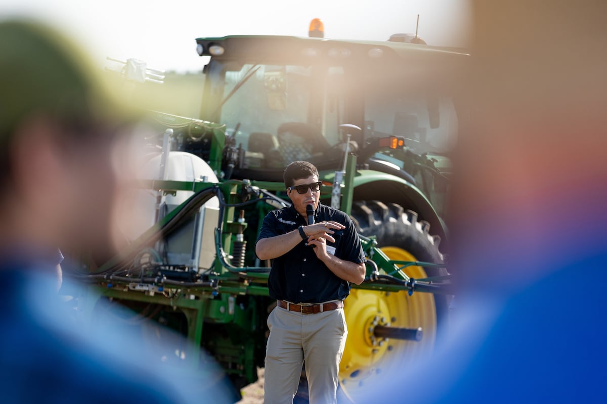 A person speaks into a microphone in front of a green tractor, with blurred foreground figures suggesting an audience.