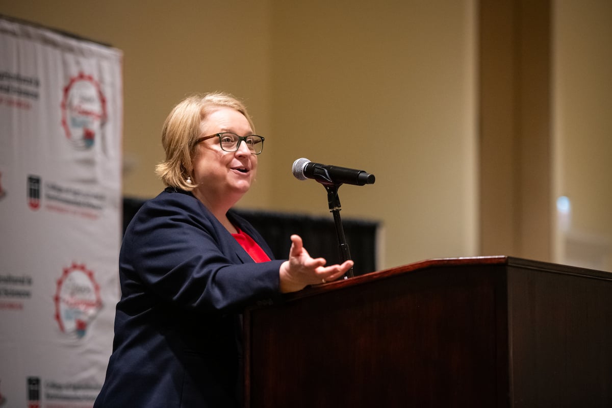 Faith Critzer gives opening remarks at a microphone in front of as branded banner
