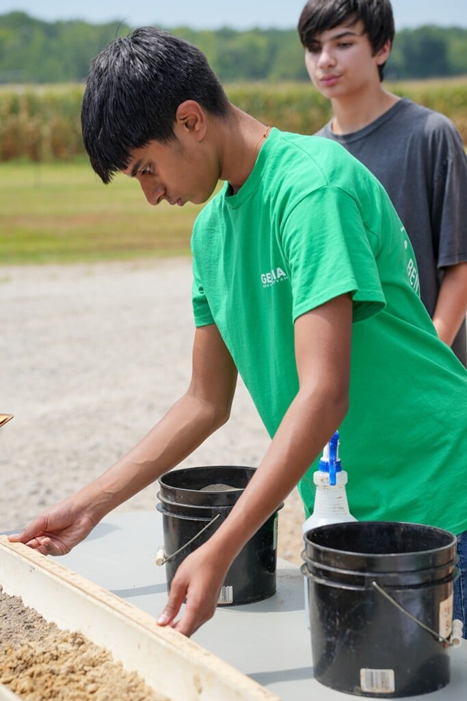 A young man in a green t-shirt examines a soil sample observed by a second young man observes