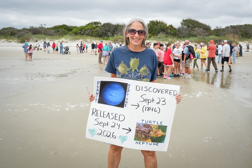 A woman stands on Jekyll Island’s beach holding a handmade poster celebrating loggerhead sea turtle Neptune’s release on Sept. 24, 2026, as visitors gather in the background.