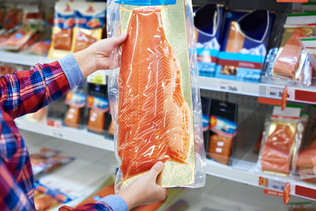 A person holding up a vacuum packed salmon filet in front of shelves of other packaged fish at a supermarket.