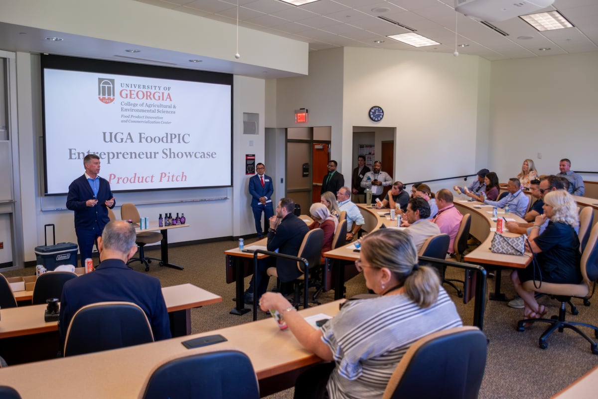 A presenter delivers a product pitch at the University of Georgia's FoodPIC Entrepreneur Showcase, with an audience seated at desks in a conference room.