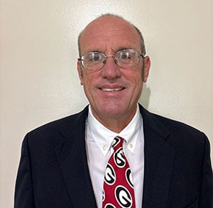 A smiling man wearing glasses and a black blazer, white shirt and red tie emblazoned with the UGA Super G.